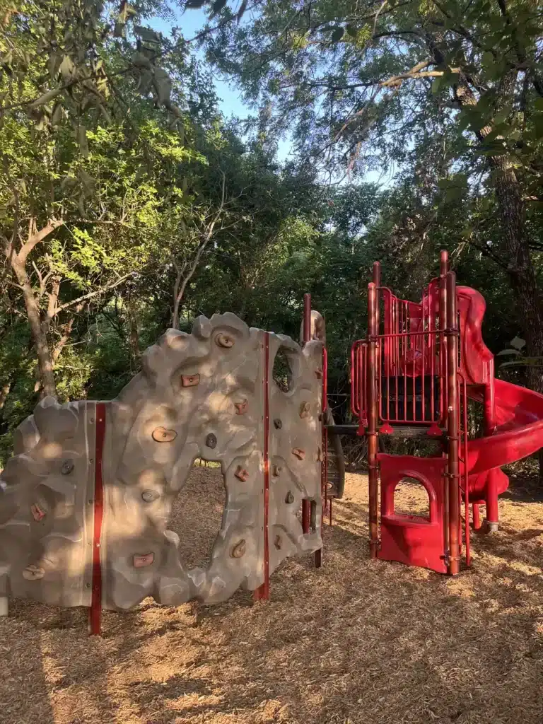 Playground with a gray molded rock climbing wall and red spiral slide tower on wood mulch beneath shady trees.